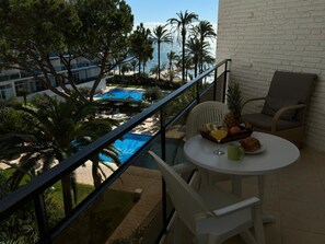 En balkong med utsikt över en pool och palmer, ett bord med en drink och frukt, och en stol.