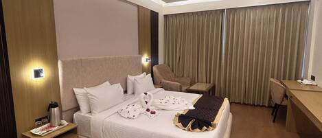 A modern hotel room with a large bed, a desk, and a chair. There is a nightstand with a lamp and a coffee maker.