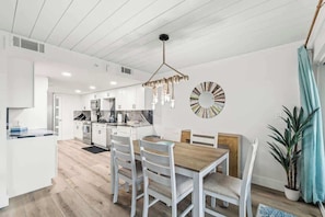 Une cuisine moderne avec des meubles blancs, un coin repas doté d’une table en bois et de chaises blanches, et un luminaire suspendu.