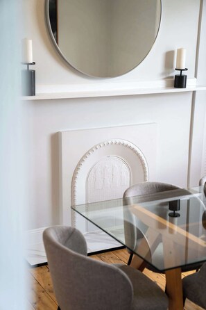 Un comedor moderno con una mesa y sillas de vidrio, una chimenea blanca con un revestimiento decorativo y un espejo redondo sobre el revestimiento.