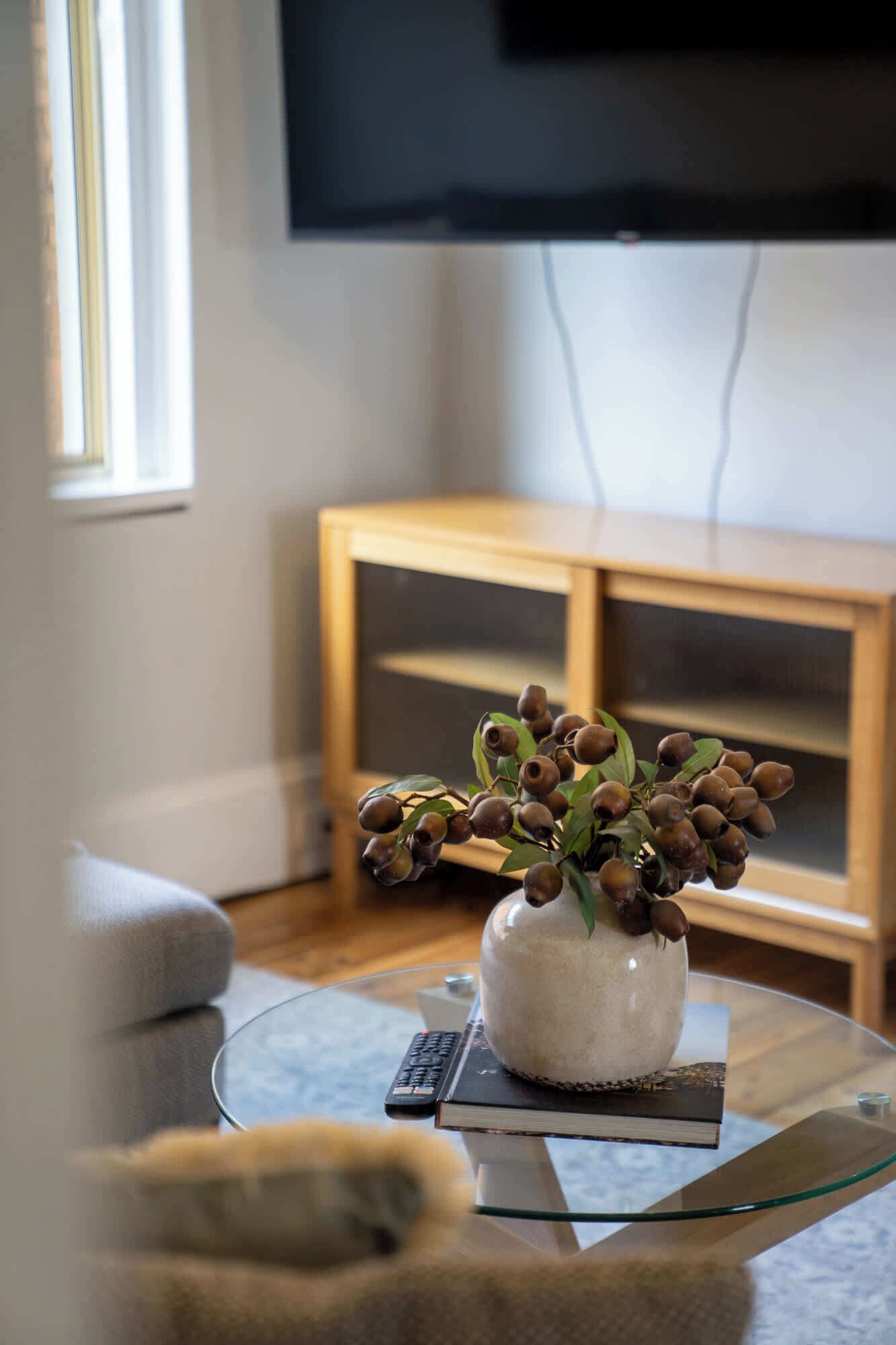 A living room with a glass coffee table, a vase with dried berries, a remote control, and a television on a stand.