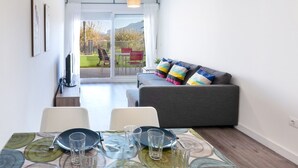 A modern living room with a grey sofa, colorful cushions, a dining table with chairs, and a large sliding glass door leading to an outdoor area with a red chair and a table. - Happy  - Apartment In Urbanització Platja D'oliva (Oliva Nova, Valencia)