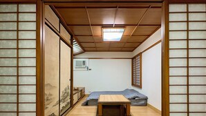 Una habitación japonesa tradicional con suelo de tatami, una mesa baja de madera, un futón como cama, puertas correderas con paneles shoji y un pequeño equipo de aire acondicionado en la pared.