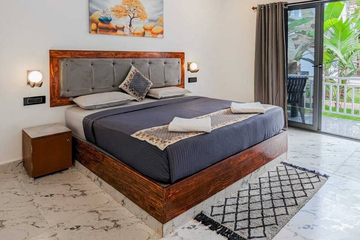 A bedroom with a large bed, a ceiling fan, a nightstand, a wall-mounted lamp, and a painting above the bed.