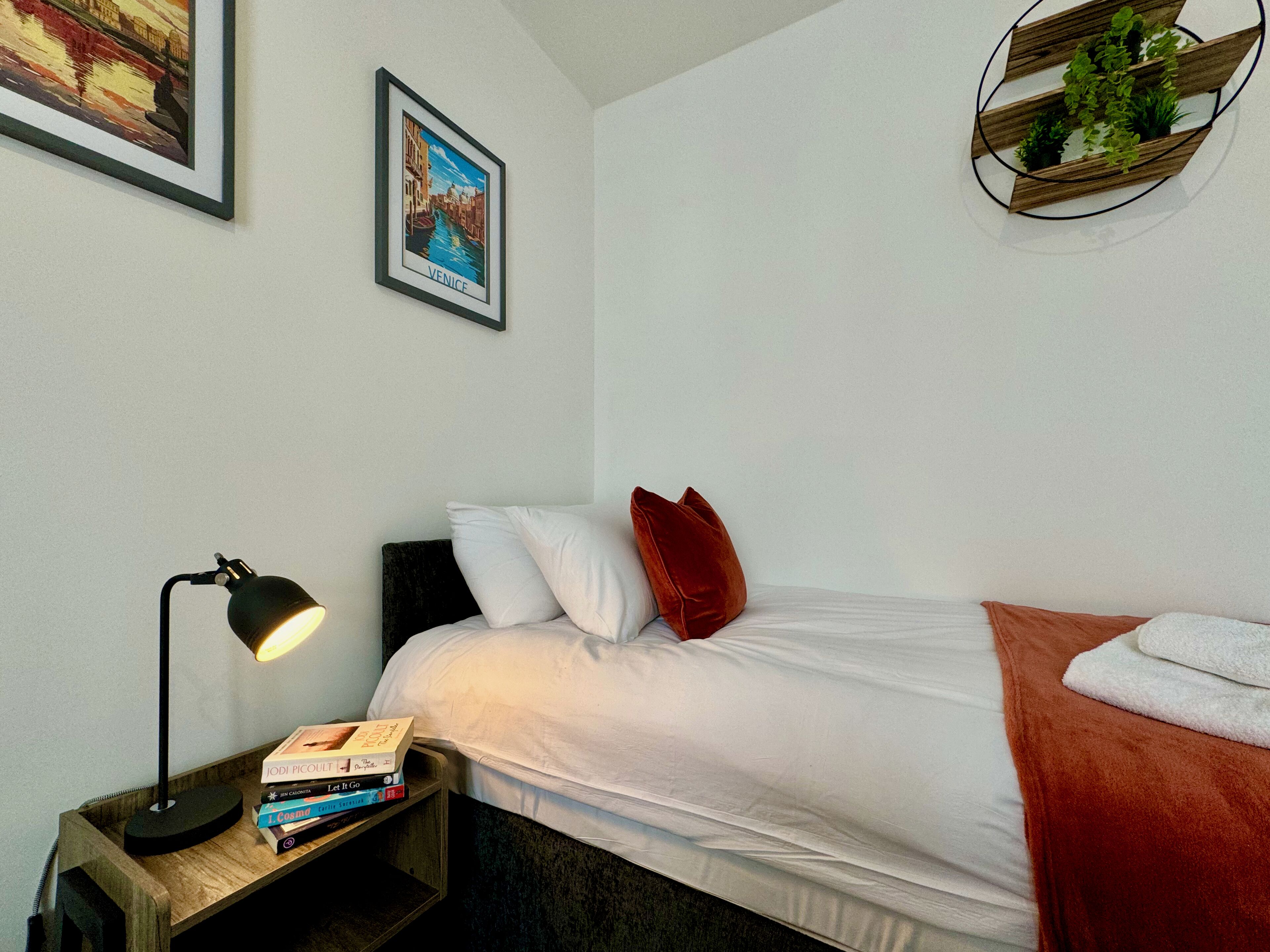 Una camera da letto ordinata con un letto, un comodino con libri, una lampada e un quadro appeso al muro.