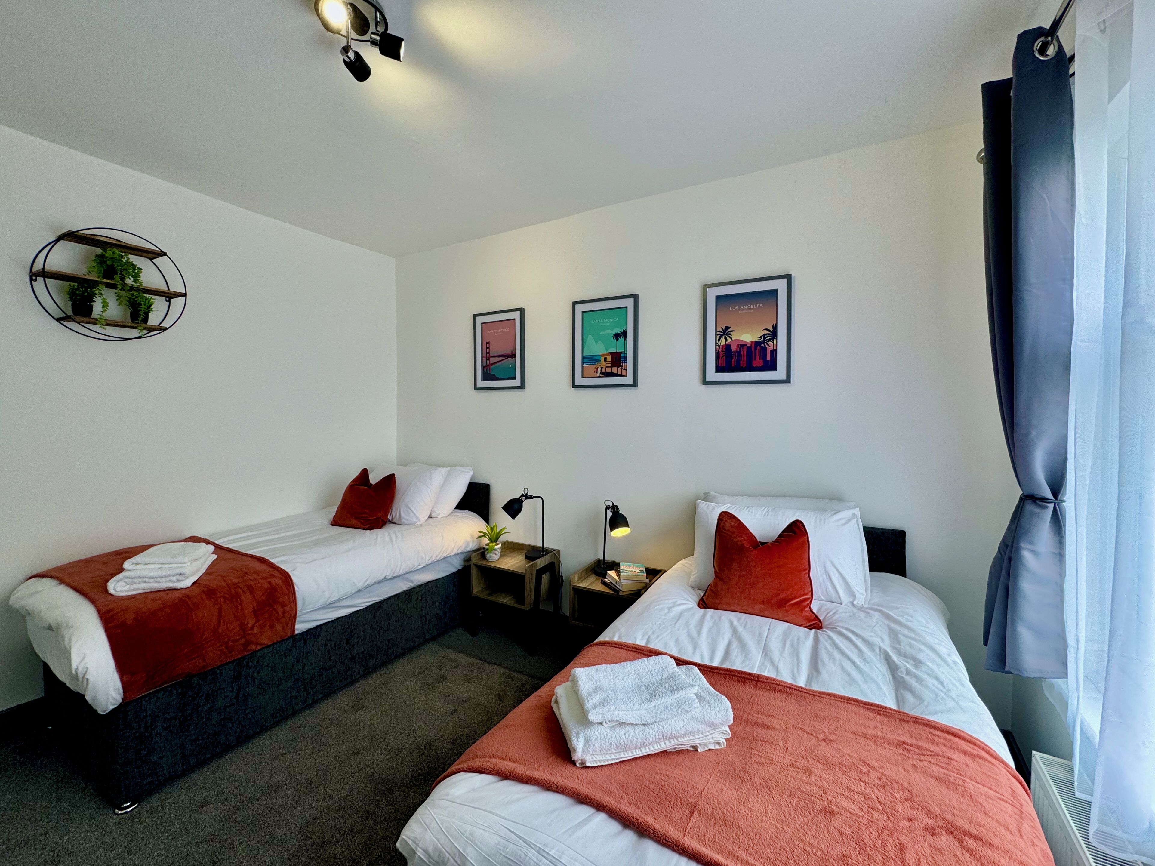 Et hotelværelse med to senge, hver med hvide sengetøj og røde puder, et lille natbord med en lampe og en vægmonteret hylde med planter.
