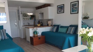 A modern, well-lit room with teal bedding, a wooden chest of drawers, a small kitchen area, and a dining table with a vase of flowers.