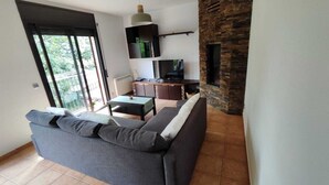 A modern living room with a grey sofa, a small table, and a TV. There is a balcony with a view of greenery outside. - RURAL APARTMENT SOL BERGUEDA (Avià Barcelona)