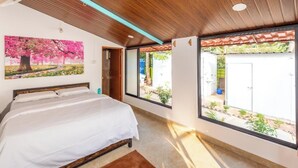 A bedroom with a bed, a ceiling fan, a large window with a view of the outside, and a painting on the wall. - Olive Beach House (Canacona)