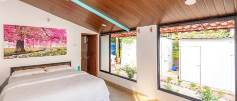 A bedroom with a bed, a ceiling fan, a large window with a view of the outside, and a painting on the wall.