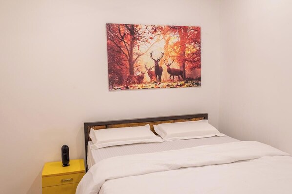 A bedroom with a bed, white bedding, a yellow bedside table with a lamp, and a wall-mounted picture of deer in a forest. - Olive Beach House (Canacona)