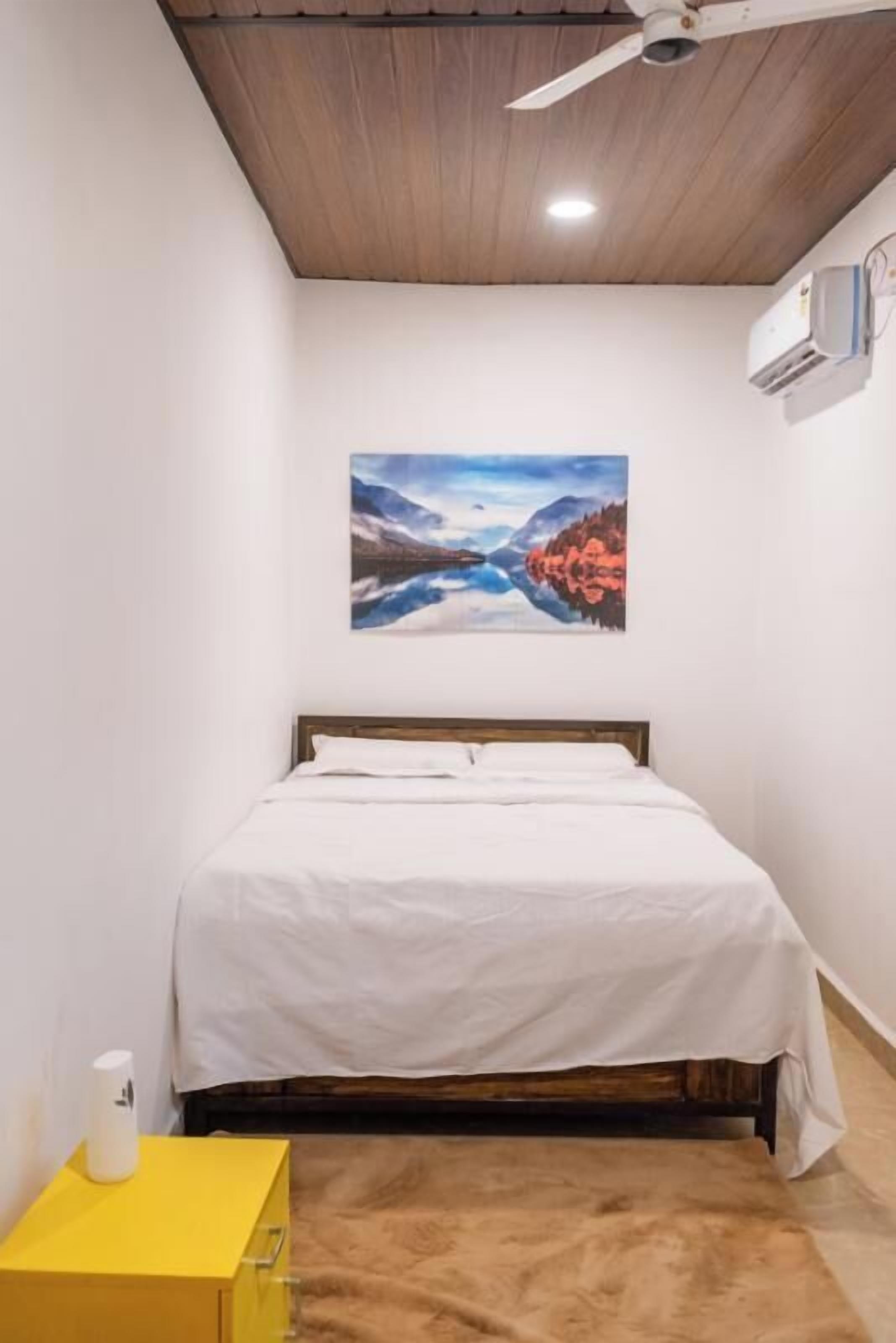 A bedroom with a bed, a ceiling fan, an air conditioning unit, a painting on the wall, and a yellow nightstand.