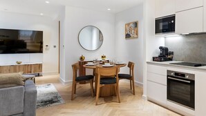 A modern living room with a grey sofa, a dining table with chairs, a flat-screen TV, a round mirror, and a kitchen with white cabinets and appliances. - Stylish Flat-Leisure Pool & Gym (Greater London)