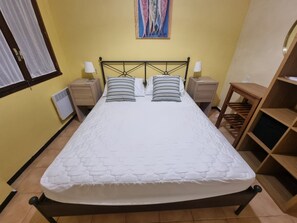 Una cama bien hecha con colcha blanca, dos almohadas, cabecera de madera y mesita de noche con lámpara.