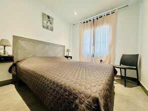 A neatly made bed with a quilted cover, a headboard, a bedside table with a lamp, a chair, and a window with curtains.