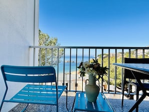 A balcony with a view of the sea, a blue chair, a table, and a vase with flowers.