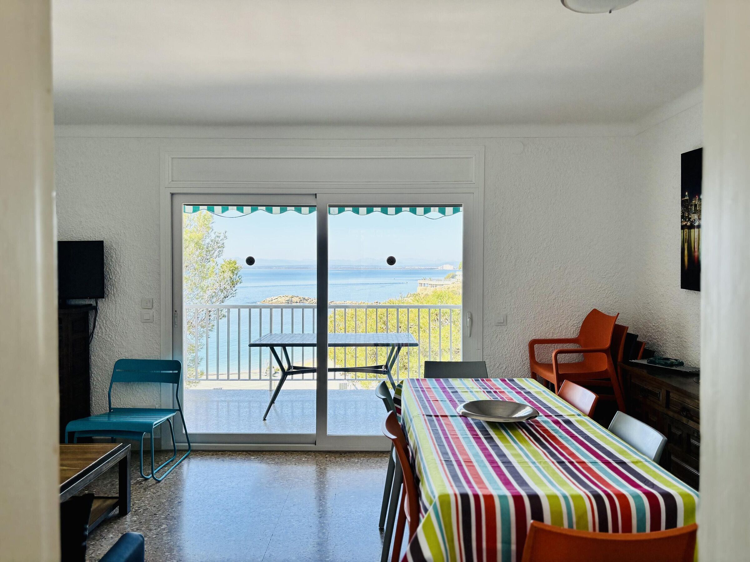 A room with a dining table, chairs, a TV, and a view of the sea through a sliding door.