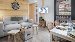 Un salon chaleureux avec un canapé gris, des murs en bois, une horloge murale, un téléviseur et un coin repas avec une table mise pour deux personnes.