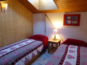 Quarto com duas camas, paredes de madeira, uma pequena mesa com abajur e um quadro na parede.