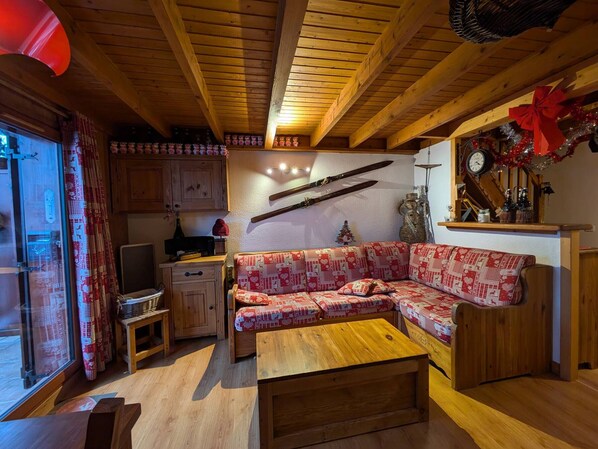A cozy living room with a wooden ceiling, a sofa with a red and white pattern, a wooden coffee table, and a kitchen area with wooden cabinets.