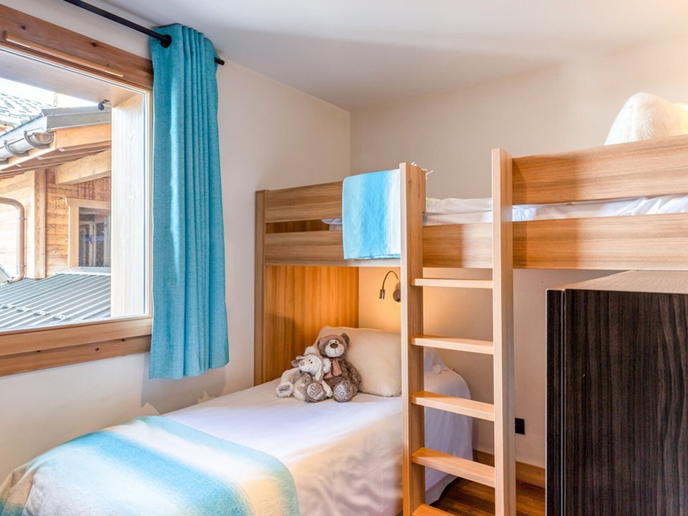 A compact bedroom with a bunk bed, a small bedside table, a teddy bear, and a window with curtains.