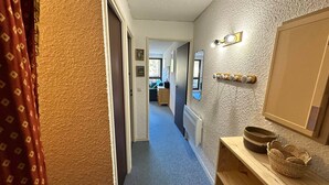 A narrow hallway with a blue carpet, a wooden shelf with a woven basket, a wall-mounted light fixture, and a door leading to another room.
