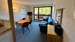 A room with a dining table, chairs, a sofa, a television, and a large window with a view of trees.
