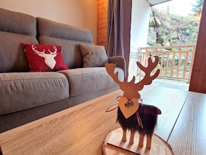 Une décoration représentant une tête de cerf en bois est posée sur une table dans un salon, où se trouvent un canapé gris et un coussin rouge orné d’un motif de cerf blanc.