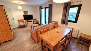 A wooden dining table with six chairs, a television, and a sofa in a room with wooden flooring and a large window. - Large apt 6/8 ppl, direct slopes access, near center, terrace (Valfréjus)