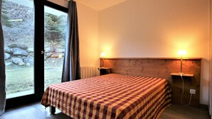 A bedroom with a checkered bedspread, two bedside lamps, a wooden headboard, and a view of a stone wall and greenery outside. - Large apt 6/8 ppl, direct slopes access, near center, terrace (Valfréjus)