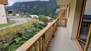 Un balcon doté d’une balustrade en bois, offrant une vue sur un paysage montagneux, ainsi qu’une zone de stationnement et des bâtiments en arrière-plan.