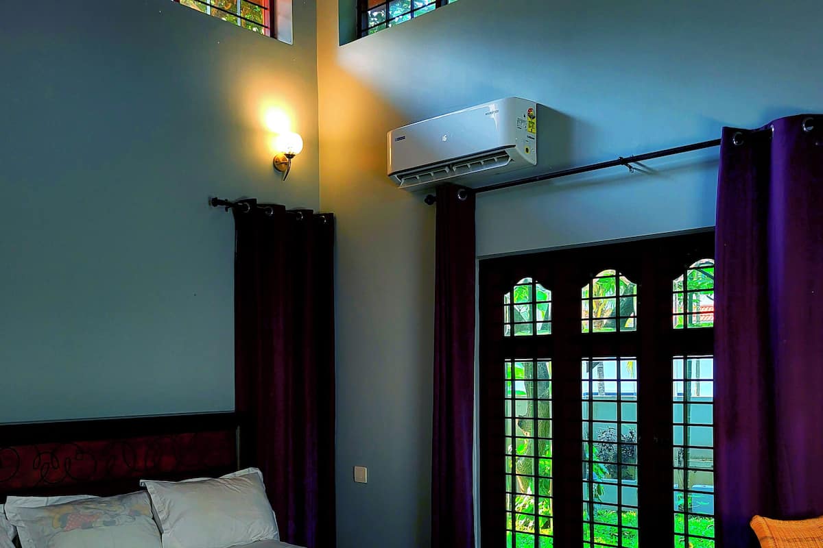 A hotel room with a bed, an air conditioning unit, a window with curtains, and a view of greenery outside.