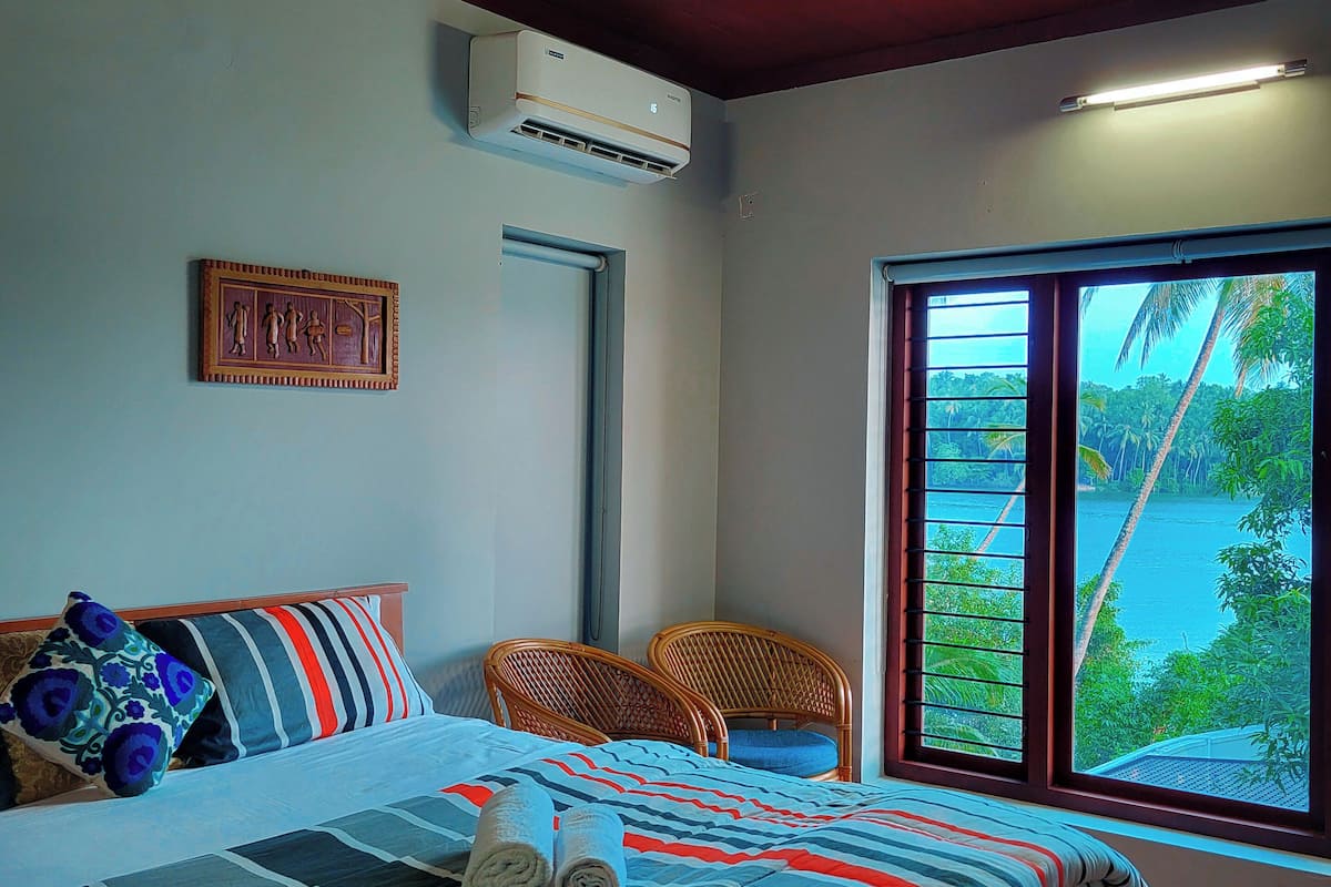 A bedroom with a bed, two chairs, a ceiling fan, an air conditioning unit, a window with a view of greenery, and a door.