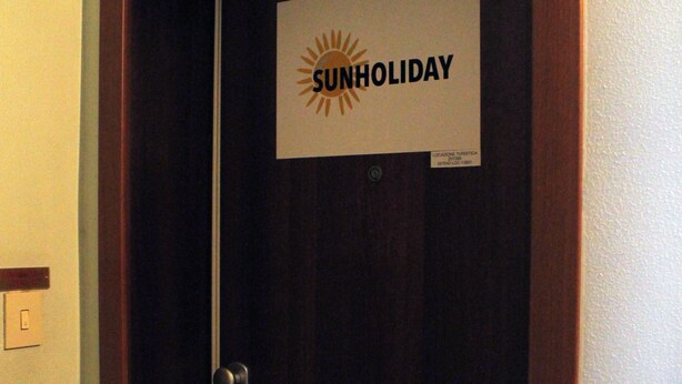 Una porta de fusta tancada amb un rètol que diu "SUNHOLIDAY" i una petita etiqueta a sota.