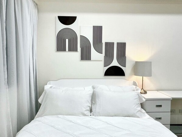 A modern bedroom with a bed, white bedding, a nightstand with a lamp, and three abstract wall art pieces.