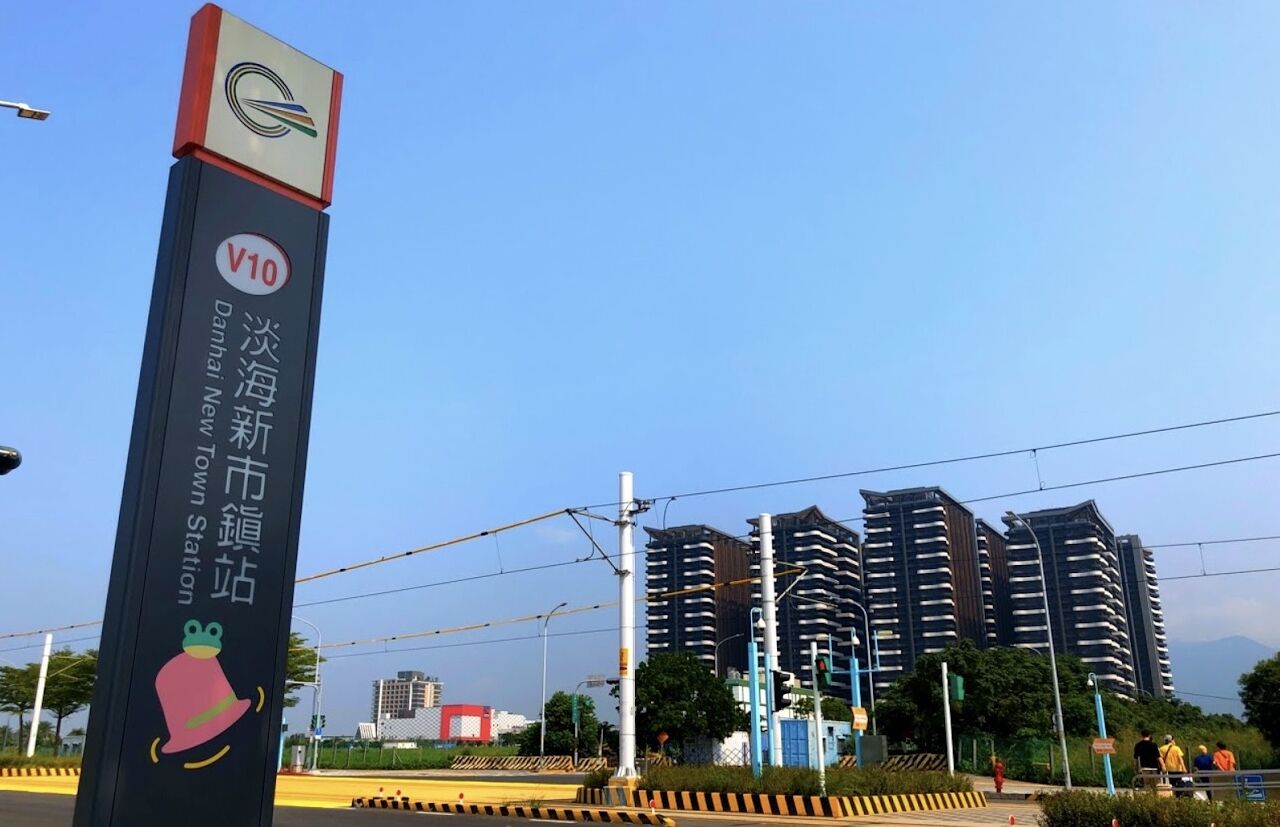 A sign for Danhai New Town Station with a V10 logo, surrounded by modern high-rise buildings and a clear sky.