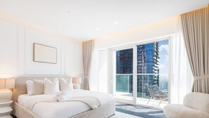 A modern hotel room with a large bed, a sitting area, and a view of a cityscape through floor-to-ceiling glass doors.