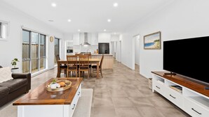 A modern living room with a sofa, a wooden coffee table, a TV stand, and a dining area with chairs and a kitchen in the background. - Point Retreat in The Point (POINT LONSDALE)