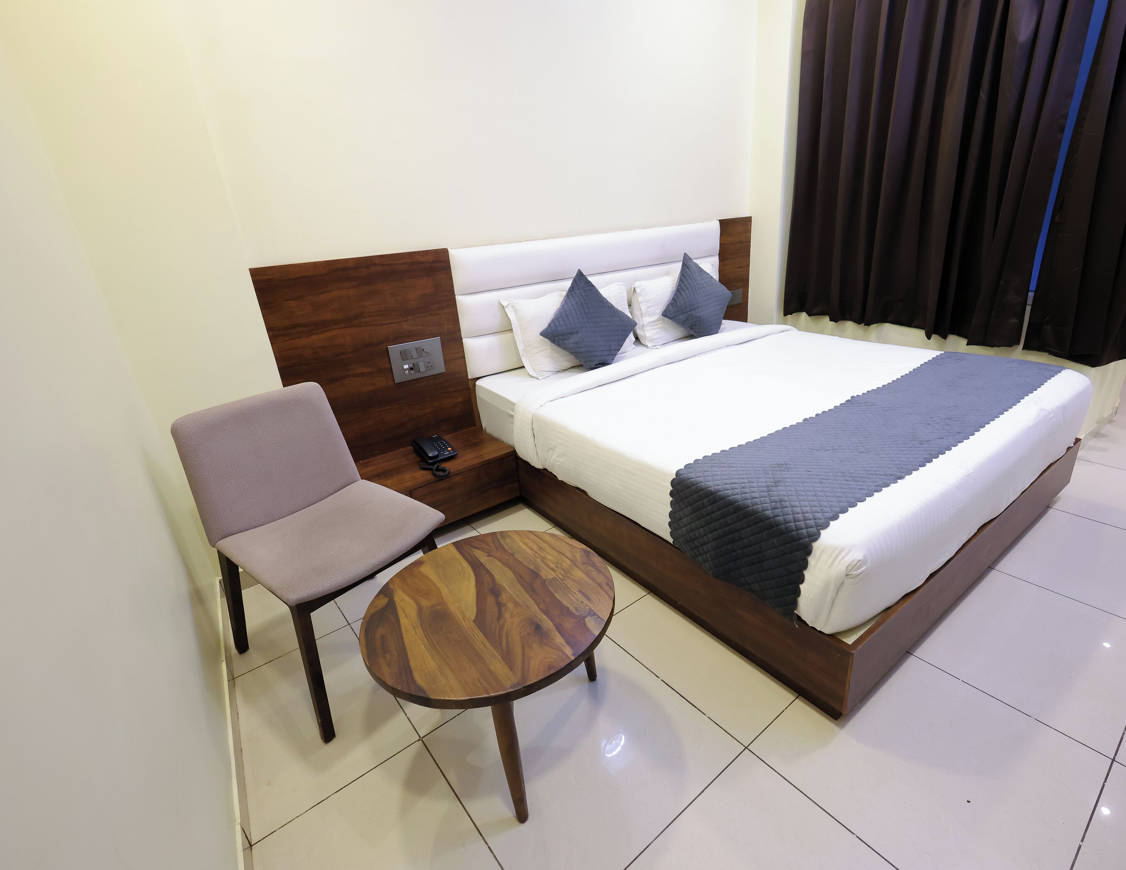 A hotel room with a bed, a chair, a small table, and a bedside table with a phone.