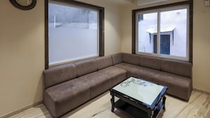 A living room with a brown sectional sofa, a glass coffee table, large windows, and a wall clock. - KANHA HOTEL (Nathdwara)
