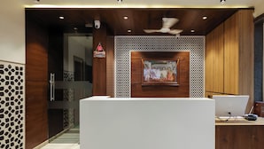 A reception desk with a white counter, wooden paneling, a ceiling fan, and a framed artwork above the counter. - KANHA HOTEL (Nathdwara)