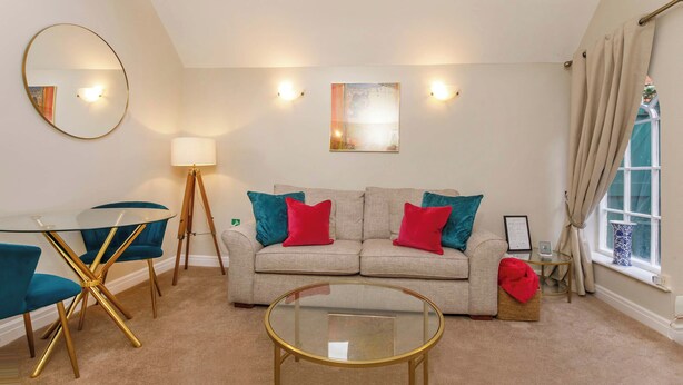 Een moderne woonkamer met een glazen salontafel, een bank met rode en turquoise kussens, een ronde spiegel en een lamp.