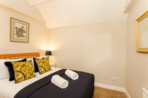 A neatly made bed with a black bedspread and two rolled white towels, two yellow pillows with floral designs, a wooden headboard, a bedside table with a lamp, and a framed picture on the wall.