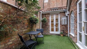 A brick building with a small patio area featuring a table and chairs, a hanging plant, and a white door. - Monk Bar Garden Hideaway (York)