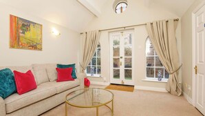 A living room with a beige sofa, red and teal cushions, a glass coffee table, and a large window with curtains. - Monk Bar Garden Hideaway (York)
