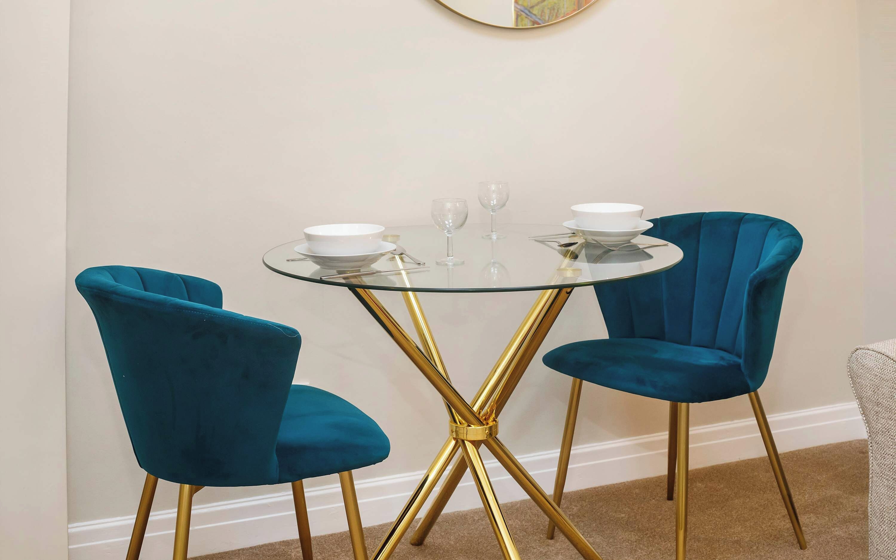 A dining area with a round glass table and two blue velvet chairs with gold legs.