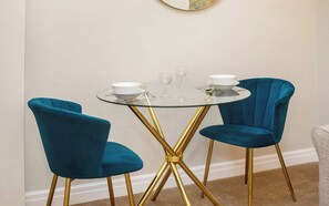 A dining area with a round glass table and two blue velvet chairs with gold legs.