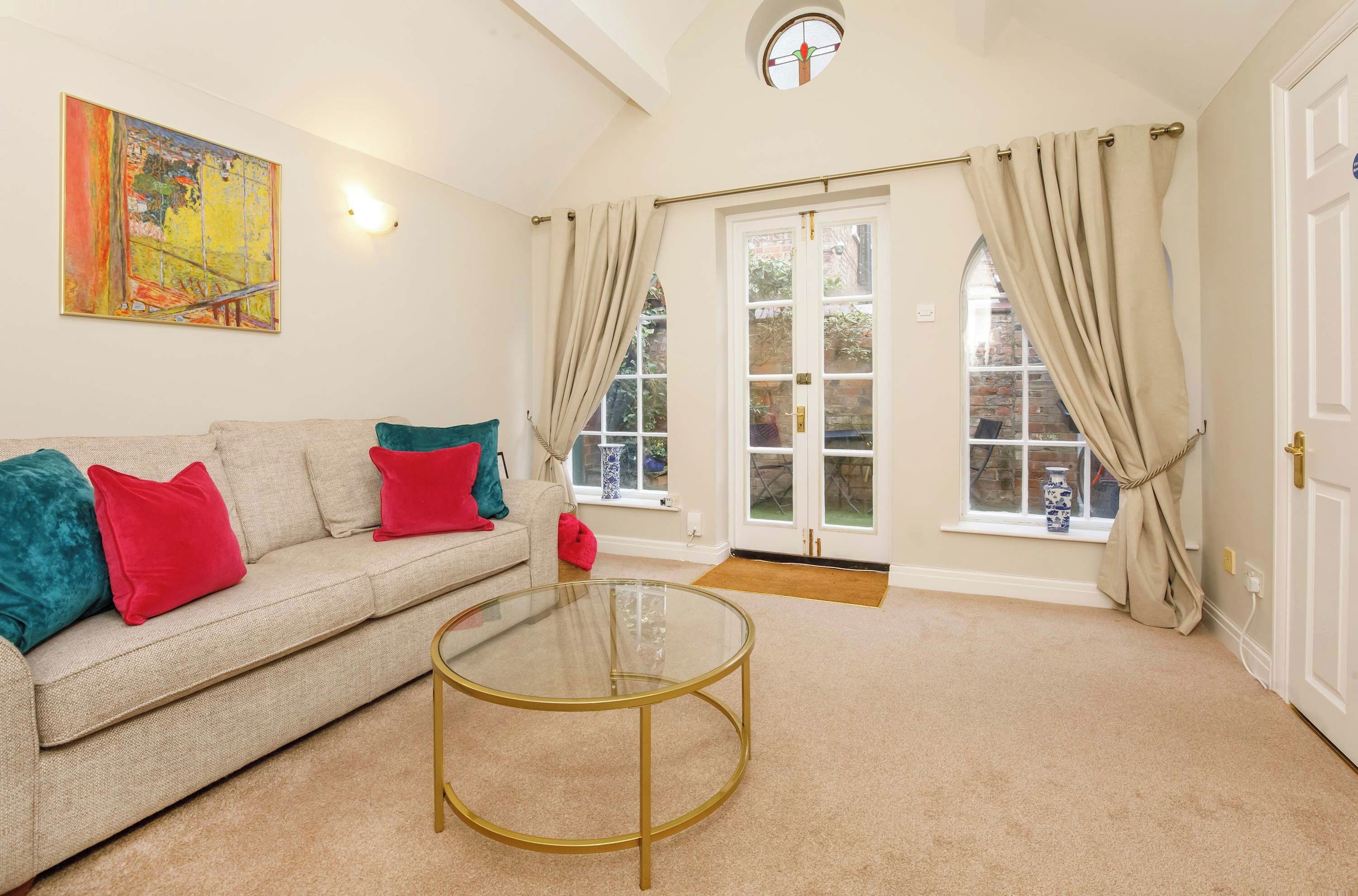 A living room with a beige sofa, red and teal cushions, a glass coffee table, and a large window with curtains.