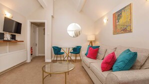 A modern living room with a glass coffee table, teal chairs, a beige sofa with colorful cushions, a flat-screen TV, and a large mirror. - Monk Bar Garden Hideaway (York)