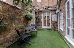 A brick building with a small patio area featuring a table and chairs, a hanging plant, and a white door.
